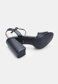 Mustat korot sandaalit, joissa on sileä nahkaosa, nilkkahihna ja lohkare korko. Teksturoitu pohja ja pyöreä varvasosa. Minimalistinen muotoilu.