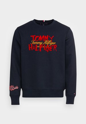 Sweat-shirt bleu marine avec inscription du logo Tommy Hilfiger en rouge et jaune sur la poitrine et texte brodé rouge sur le poignet de la manche gauche.