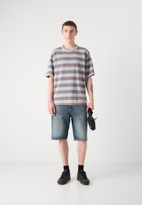 Grå og brun stribet t-shirt, løs pasform, med denim shorts. Sorte sneakers og holder en lille sort taske. Ensfarvet hvid baggrund.