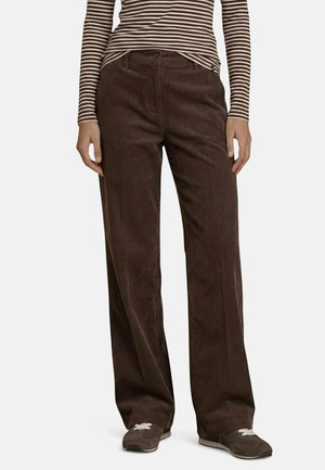 Personne portant un pantalon large en velours côtelé marron, une chemise à manches longues rayée brun et blanc, et des baskets marron avec des lacets blancs.