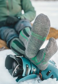 Calcetines grises con acentos verdes, con el texto "Danish Design", combinados con fijaciones de esquí turquesa sobre un fondo de nieve blanca.