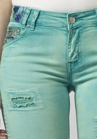 Denim traperice u akvablu boji s izrezbarenim detaljima i dekorativnim šljokicama na prednjem džepu. Imaju standardno kopčanje na dugme i zatvarač.
