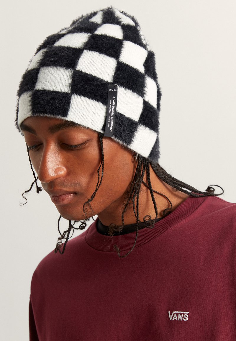 Černobílá kostkovaná pletená čepice s nadýchanou texturou a značkovým štítkem po straně, kterou nosí osoba v vínovém svetru.