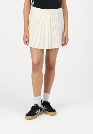 Pleated mini skirt in cream fabric with a smooth texture, featuring a wide waistband and a hem just above the knee. Paired with black sneakers.