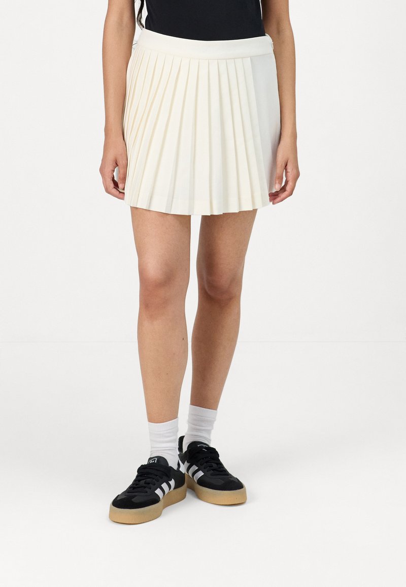 Pleated mini skirt in cream fabric with a smooth texture, featuring a wide waistband and a hem just above the knee. Paired with black sneakers.