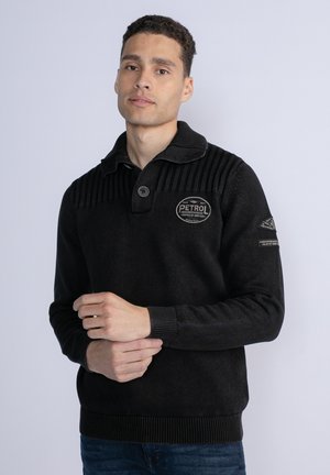 Black knit sweater with a ribbed collar, button closure, and logo patch on the chest. Features textured details on the shoulders and cuffs.