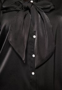Blusa nera in satin con un grande collo a fiocco. Caratterizzata da bottoni simili a perle e una texture liscia. Design classico con dettagli eleganti.