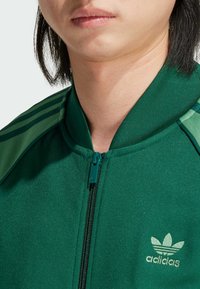 Zelená bunda na zip s texturovanou tkaninou, s tmavšími zelenými pruhy a malým logem adidas ve světle zelené barvě na levé hrudi.