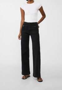 Vaqueros negros de pierna ancha con cintura alta, bolsillos delanteros y detalles en botones plateados, combinados con una camiseta blanca ajustada de manga corta.