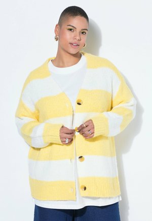 Persona che allaccia un cardigan a righe gialle e bianche sopra una camicia bianca, indossando orecchini a cerchio dorati e diversi anelli, su uno sfondo semplice.