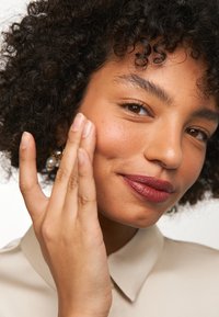 Modèle avec des cheveux noirs bouclés et des boucles d'oreilles en or. Elle porte une chemise beige à col, se touchant la joue avec un blush rose clair et un rouge à lèvres foncé.