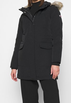 Personne portant un parka d'hiver noir avec capuche bordée de fourrure, fermeture éclair sur le devant et boutons-pression, poches à rabat et pantalon noir.