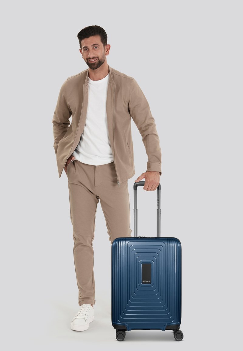 Valigia rigida di colore blu navy con un motivo a onde testurizzate, dotata di maniglia retrattile e quattro ruote, posizionata accanto a un uomo in abbigliamento casual.