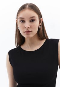 Svart ribbad ärmlös topp, slät textur, med rund halsringning. Modellen bär cirkulära silverörhängen. Minimalistisk design, neutral bakgrund.