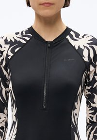 Žena oblečená v čierno-bielom vzorovanom neoprénovom tričku s dlhým rukávom na zips s logom "Billabong" na hrudi.