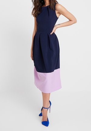 Femme portant une robe midi sans manches bleu marine et rose clair avec des plis, associée à des chaussures à talons bleues avec bride à la cheville, debout sur fond blanc.