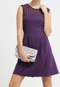Robe violette avec un décolleté en dentelle texturé, corsage ajusté, jupe évasée et une pochette en argent avec des embellissements cloutés.