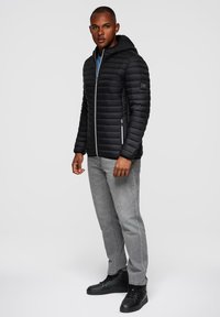 Černá péřová bunda s kapucí, prošívaný design, přední zip, boční kapsy a detail s nášivkou. Nositá s šedými džínami a černými teniskami.
