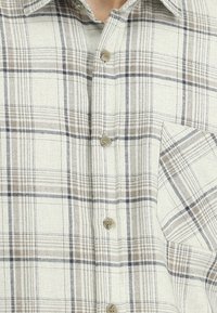 Gładka, w kratę koszula rozpinana w jasnym beżu z szarymi i brązowymi akcentami. Posiada kieszeń na piersi oraz teksturowany materiał. Zapięcie na sześć guzików.