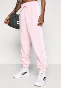 Rozā sporta bikses ar baltām sānu joslām un elastīgiem beidzējiem, kombinētas ar baltām kedām ar melniem akcentiem un mazu melnu somiņu.