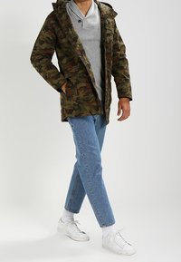 Parka s camouflage vzorem v odstínech zelené a hnědé s kapucí, nošená přes šedou mikinu, spárovaná se světle modrými džíny a bílými teniskami.