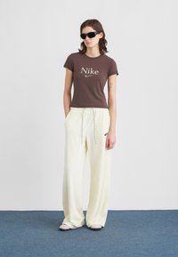 Sötét napszemüveget viselő nő barna Nike pólót és laza krém színű Nike melegítőnadrágot visel, kék szőnyegen áll egy fehér fal előtt.