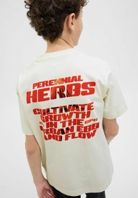 Grietos medvilnės t-shirt'as su dideliais raudonais grafikais ant nugaros, užrašytais „PERENNIAL HERBS“ ir „CULTIVATE GROWTH IN THE URBAN EBB AND FLOW.“
