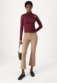 Haut à manches longues bourgogne avec découpe sur le côté et détails froncés associé à un pantalon beige évasé et des mocassins noirs ; tenant un sac vert.