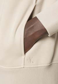 Felpa beige con polsini a coste e logo ricamato 'cK' sull'orlo. Presenta una tasca laterale, che mette in mostra una texture liscia e morbida.