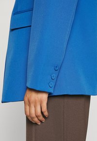 Tailored blue blazer with a smooth texture, three buttons on the cuff, and a classic pocket design, paired with brown trousers.