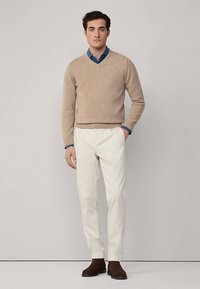 Beiger V-Ausschnitt-Pullover, hellblaues Hemd darunter, weiße Hose und braune Schuhe. Weiche Textur, tailliertes Design mit Lagen-Look.