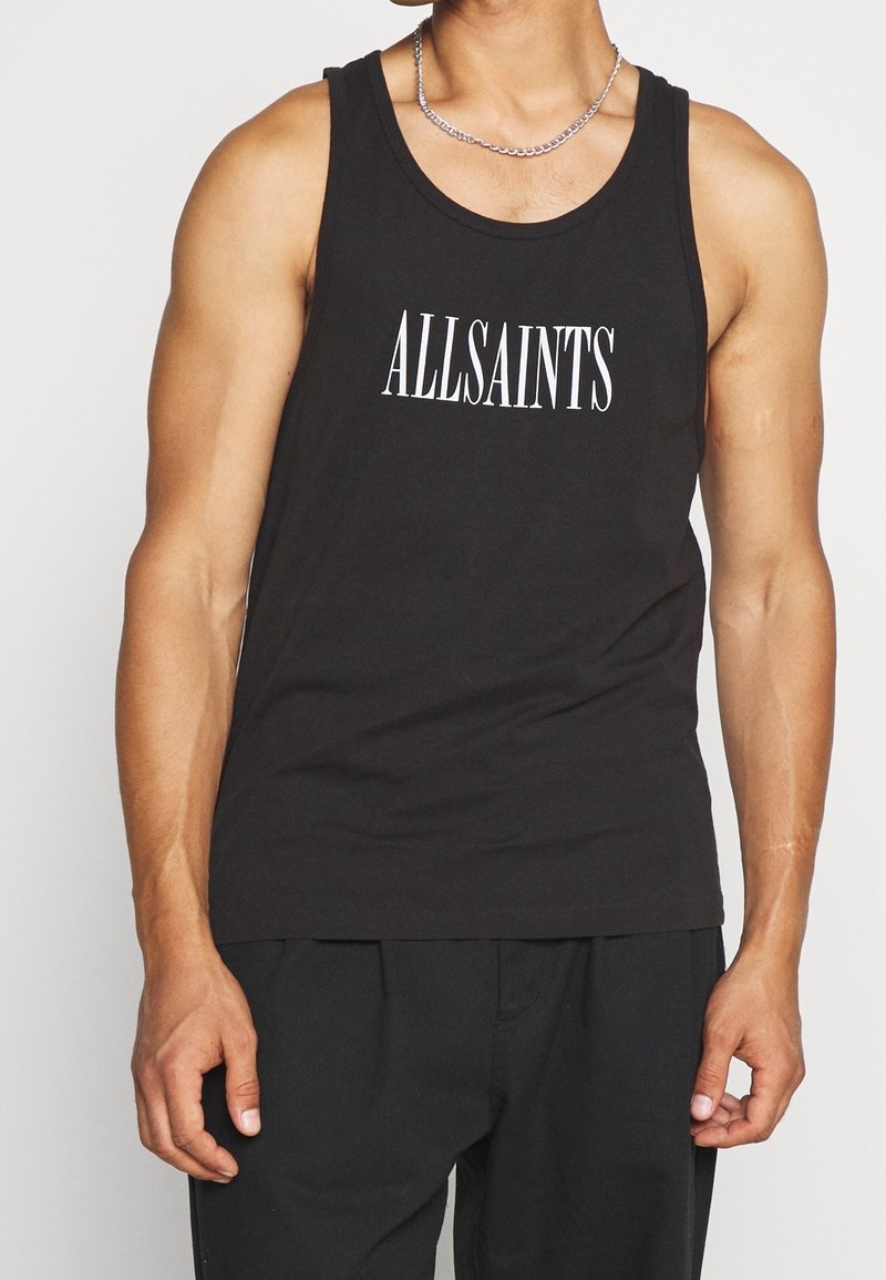 Homme portant un débardeur noir sans manches avec le texte blanc "ALLSAINTS" et un collier chaîne en argent, debout les bras détendus le long du corps.