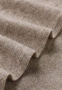 Tessuto in un morbido colore marrone chiaro, con un motivo intrecciato e testurizzato, che mostra leggere ondulazioni e pieghe.