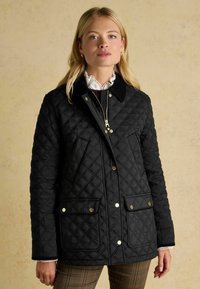 Jachetă neagră matlasată, cu guler din catifea, detalii aurii și două buzunare frontale. Designul include un model în diamante și închidere cu fermoar.