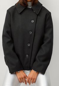 Manteau en mélange de laine noir avec un large col, cinq boutons noirs et une silhouette carrée. Les manches sont légèrement surdimensionnées et d'une texture lisse.