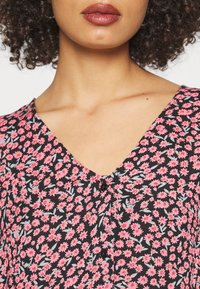 Květinová blůza z černé látky s růžovými a světle modrými květinovými vzory, ve výstřihu do V a s knoflíkovým detailem na hrudi.