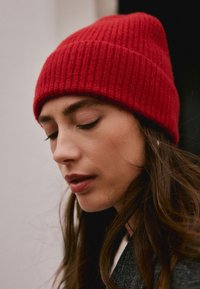 Red ribbed beanie hat made of knitted fabric, fitting snugly. Accented by a textured pattern, complementing medium-length brown hair.