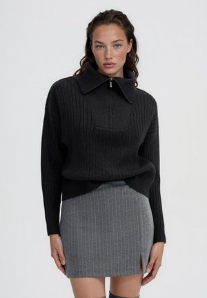 Giovane donna che indossa un maglione nero a coste con colletto con zip e una minigonna grigia a righe sottili con spacco davanti su uno sfondo semplice.