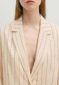 Beige gestreepte blazer met een diepe V-hals, voorzien van verticale zwarte lijnen, een enkele borstzak en een gestructureerd kraagontwerp.