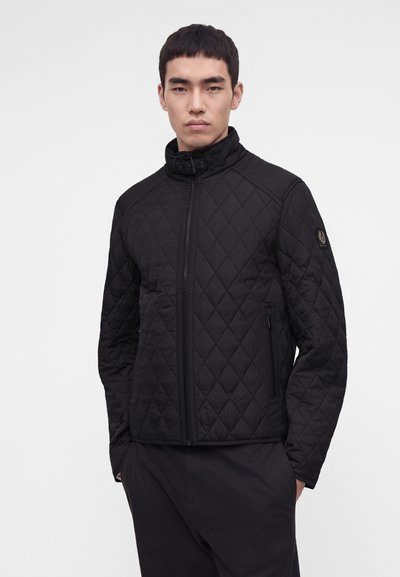 Jeune homme portant une veste matelassée noire avec une fermeture éclair à l'avant et un col montant, debout devant un fond uni clair.