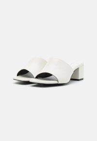 White leather block-heeled sandals with an open toe and wide strap design. Smooth texture with a matte finish and black accents on the sole.