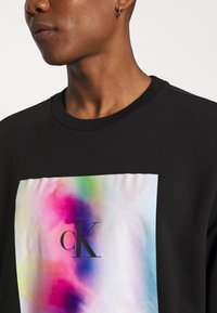 Sudadera negra con un gran графico en degradado multicolor que presenta el logo "CK" en el centro. Tela texturizada con un cuello y puños acanalados.
