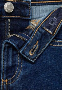 Tummansininen farkkukangas, jossa on kontrastiväriset keltaiset tikit. Siinä on metallinen nappi, vetoketju ja kangassilmukat vyön kiinnittämistä varten.