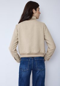 Veste bomber beige texturée avec un ourlet et des poignets côtelés, ornée de rayures blanches. Portée avec un jean denim bleu foncé.