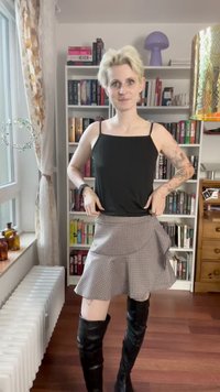 Top preto de alças finas com design justo, combinado com uma saia envolvente xadrez cinza. A modelo usa botas pretas até o joelho, exibindo tatuagens.