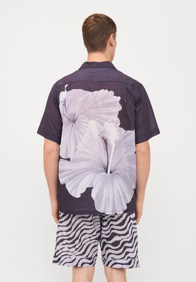 Camicia a maniche corte in blu scuro con ampio motivo floreale in bianco di hibiscus. Abbinata a pantaloni corti a fantasia zebra in bianco e nero.