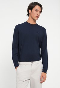 Cămașă cu mânecă lungă de culoare bleumarin, din bumbac, cu guler rotund și un mic logo în roșu, alb și albastru pe partea stângă a pieptului. Textură netedă.