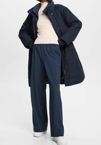 Abrigo acolchado azul marino con un cuello alto, top beige ajustado de canalé y pantalones holgados azul marino. La modelo lleva zapatillas blancas.