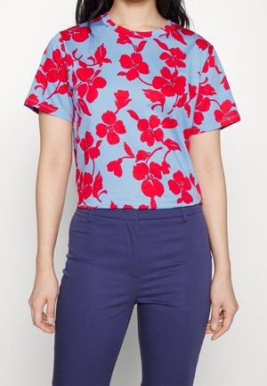 Kobieta nosząca niebieską koszulkę z krótkim rękawem z jasnoczerwonym kwiatowym nadrukiem, włożoną do ciemnoniebieskich dopasowanych spodni.