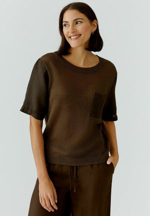 Lächende Frau mit dunklem Bob-Haarschnitt, die ein braunes kurzärmeliges Strickoberteil mit Brusttasche und passende braune Hose trägt, die Hand in der Tasche.